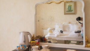A room with a bed, a white kettle, and a mirror reflecting the bed and a framed picture on the wall.
