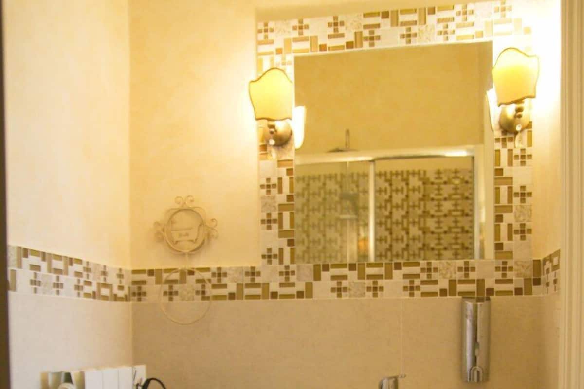 Casa de banho com lavatório branco, sanita e um pequeno banquinho branco. Há um espelho sobre o lavatório e um radiador no lado esquerdo.