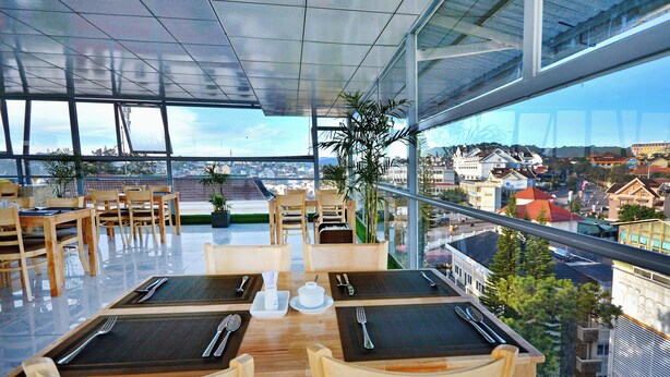 A restaurant with tables set for dining, large windows offering a city view, and indoor plants. - ELC Luxury Da Lat Hotel & Apartment (Xuan Huong - Da Lat)