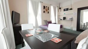 Une chambre d’hôtel équipée d’une table à manger, de chaises, d’un téléviseur, d’un lit avec des oreillers, d’une fenêtre avec des rideaux et d’une étagère fixée au mur, sur laquelle sont posés divers objets.