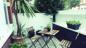 Une petite terrasse extérieure avec une table et des chaises en bois, entourée de plantes en pot et d’un banc.