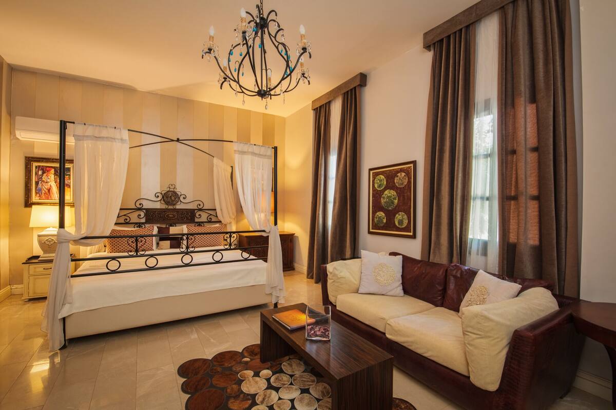 A bedroom with a four-poster bed, a sitting area with a sofa, a chandelier, and a painting on the wall.