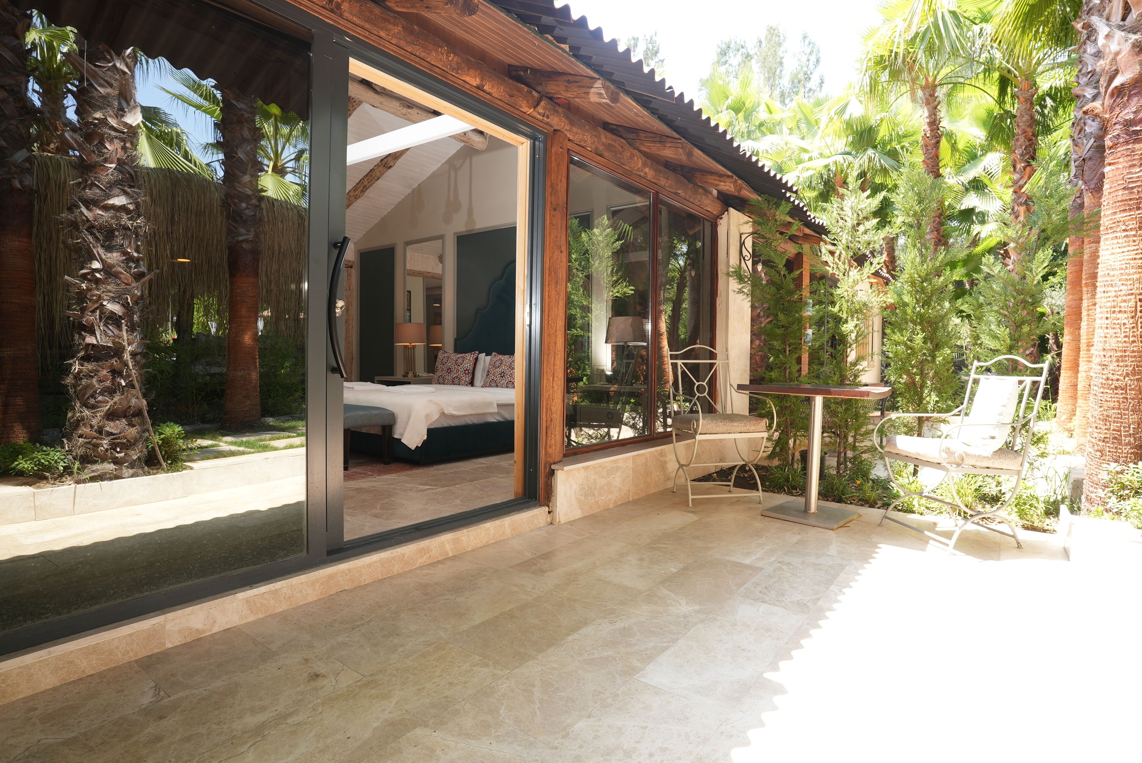 Una villa moderna con un dormitorio envuelto en vidrio, un patio con mesa y sillas, y exuberante vegetación tropical.