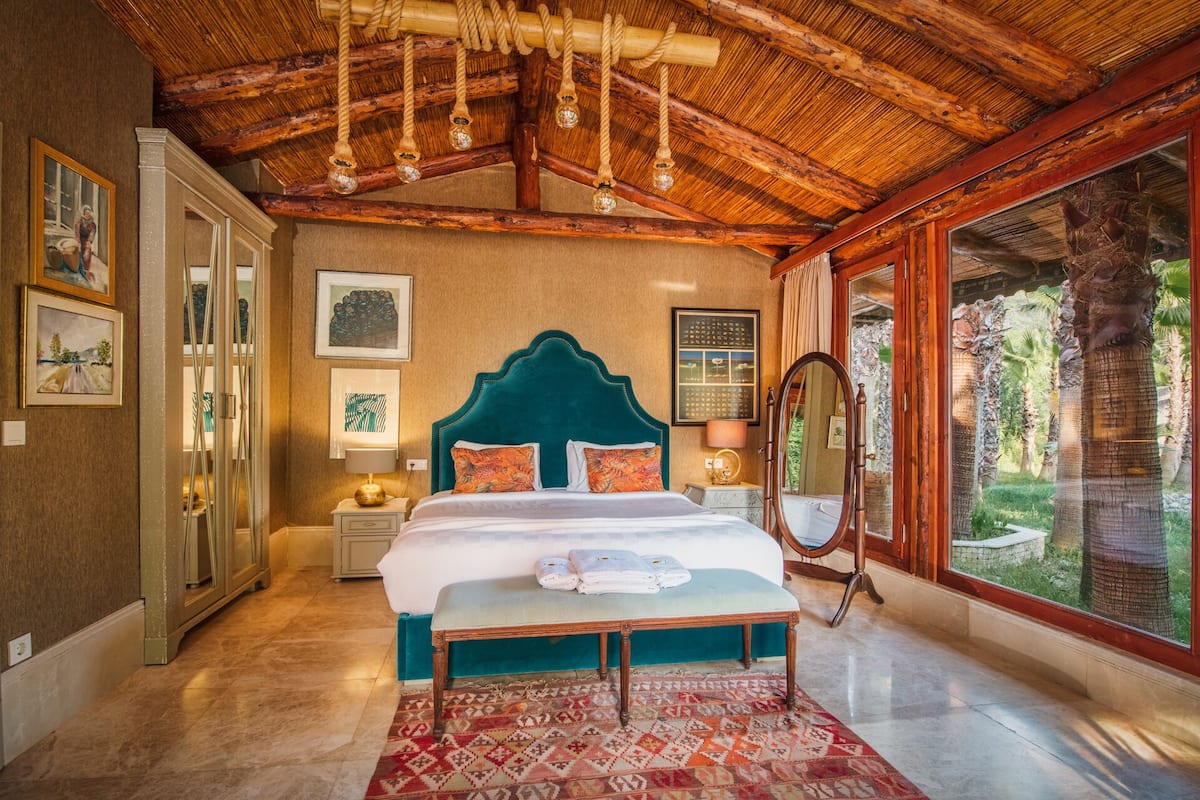 A spacious bedroom with a large bed, a wooden ceiling, and a view of a tropical garden.