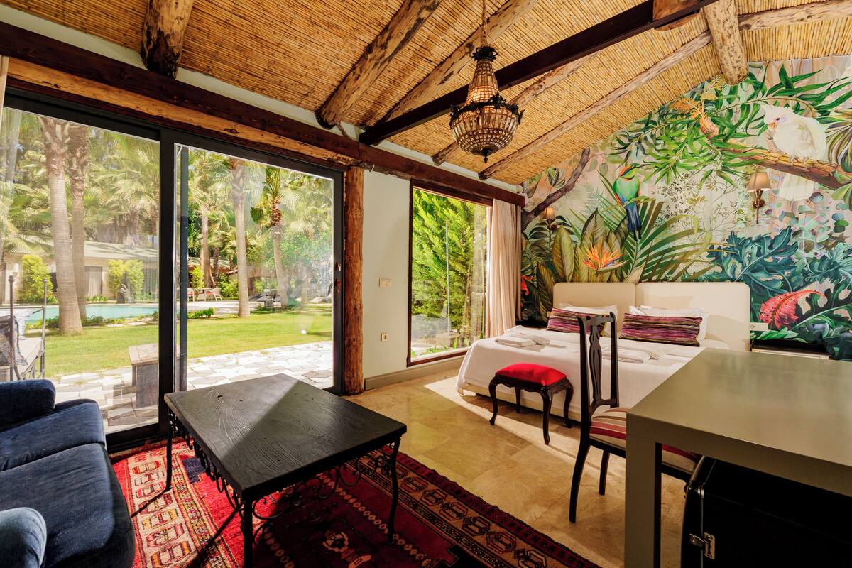 A room with a bed, a dining table, and a view of a garden and pool.