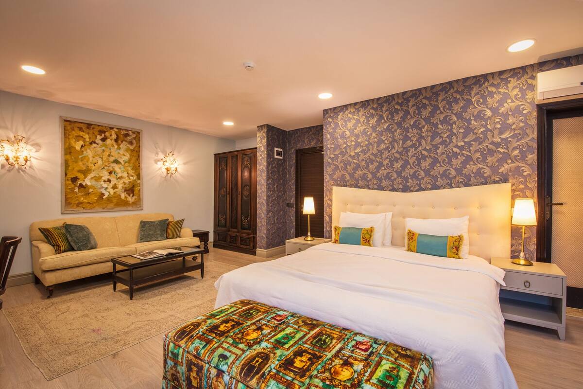 A hotel room with a large bed, a sofa, a coffee table, and a patterned ottoman.