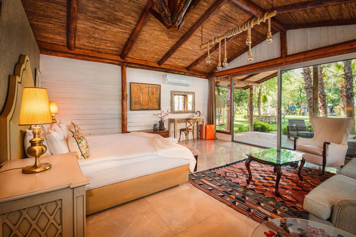 A spacious bedroom with a large bed, a desk, and a view of the garden through large windows.