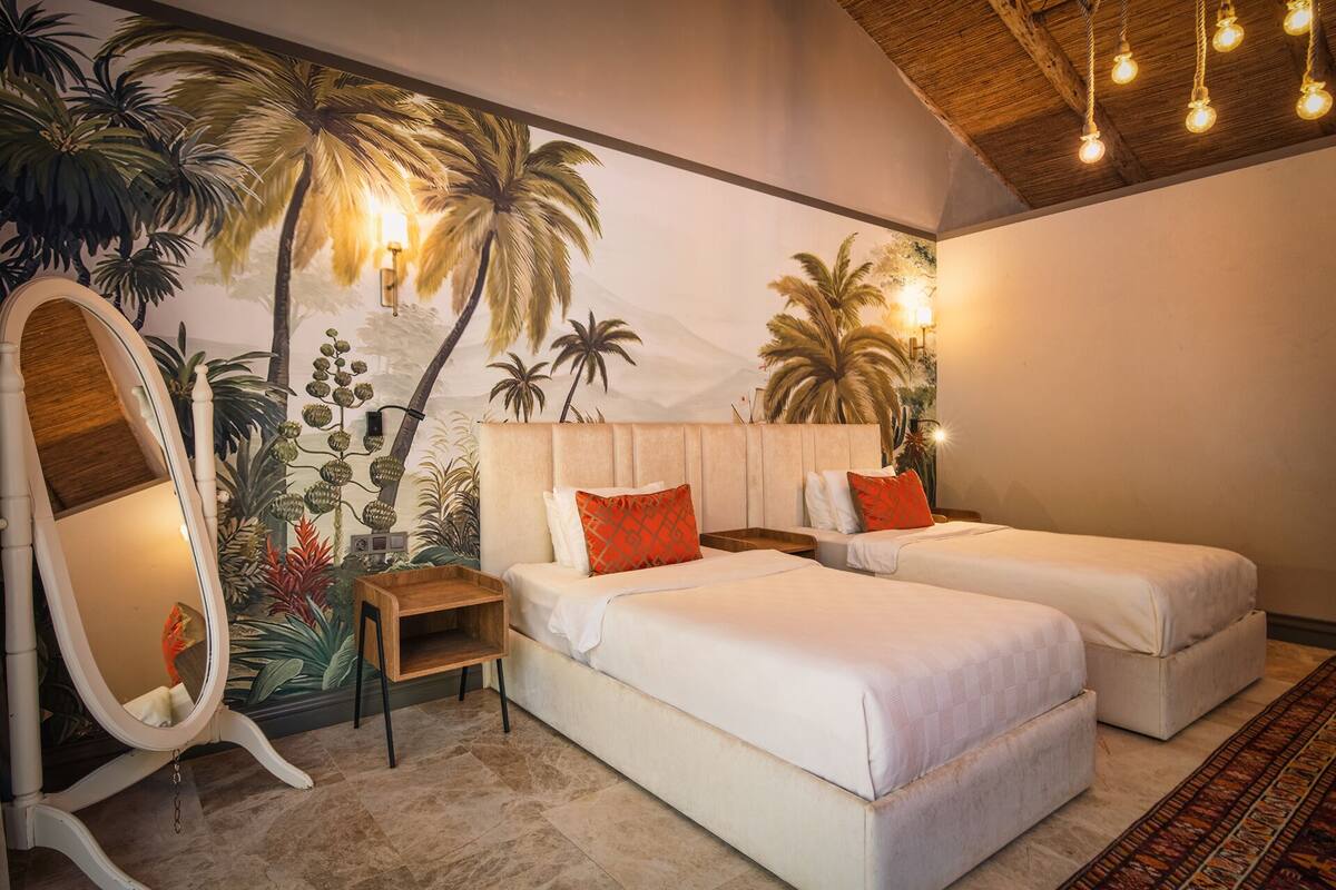 A bedroom with two beds, a large mirror, and a mural of tropical plants.