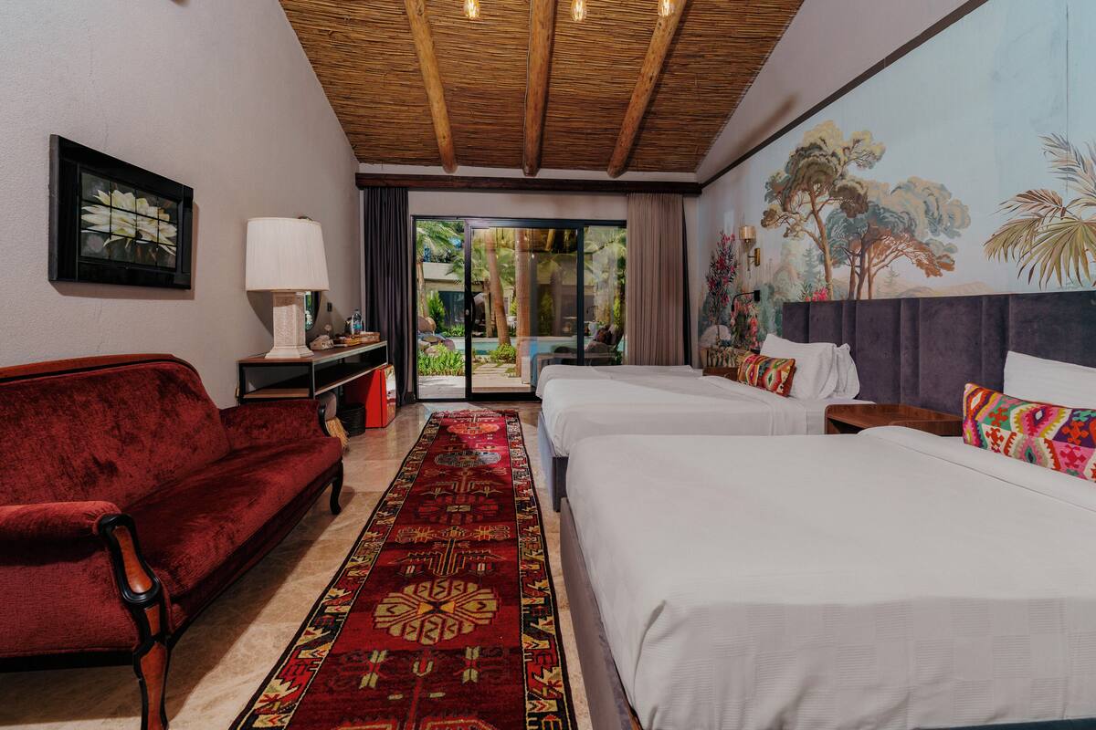 A hotel room with a red sofa, two beds, a large window, and a mural of trees.
