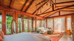 Une pièce spacieuse avec un grand lit, un canapé et une petite table. De grandes fenêtres sont ornées de rideaux, le plafond est en bois et la pièce offre une vue sur la verdure environnante.
