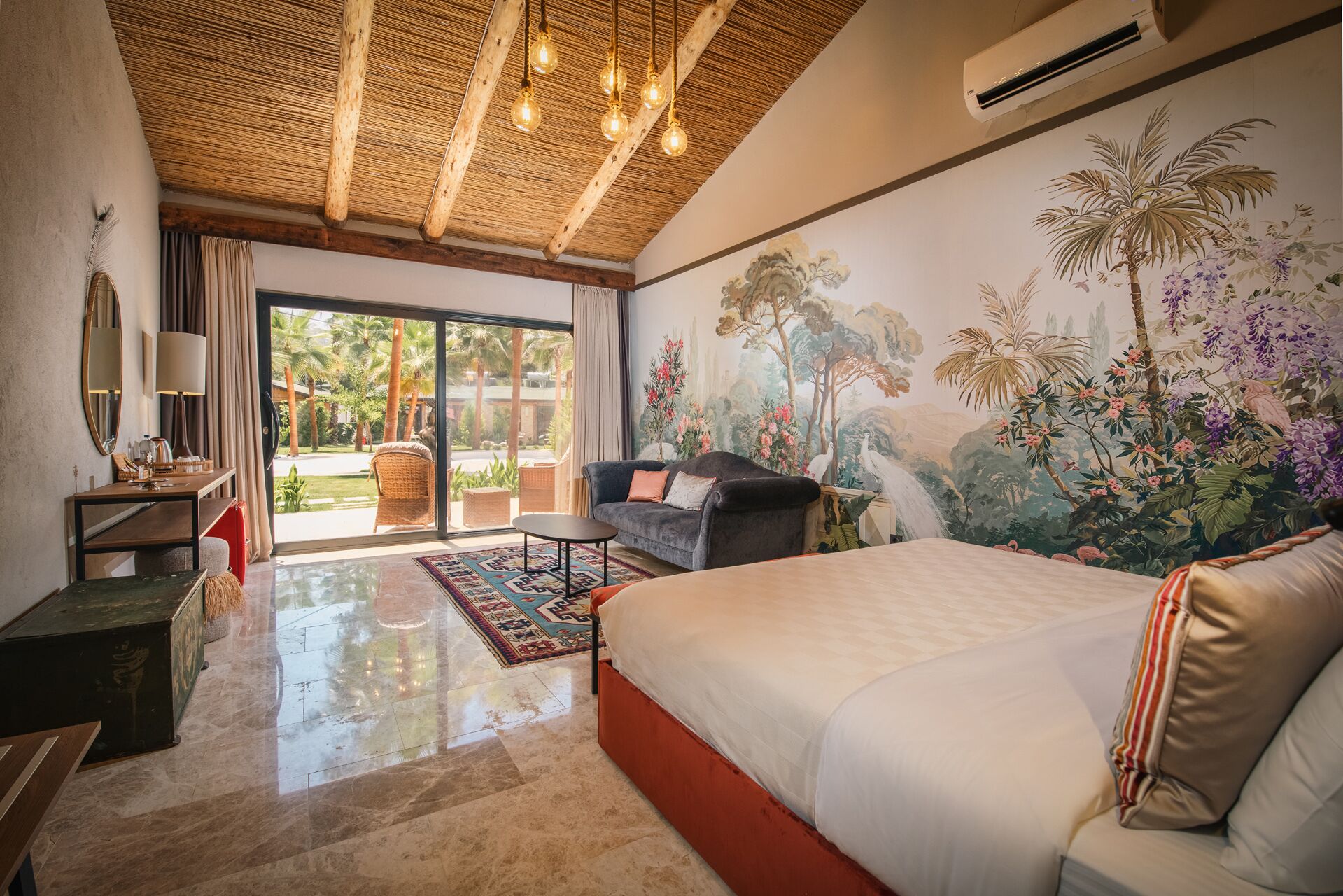 Un dormitorio amplio con una cama grande, una zona de estar con un sofá y sillones, una mesa de comedor y un mural de paisaje tropical.