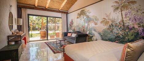 Un dormitorio amplio con una cama grande, una zona de estar con un sofá y sillones, una mesa de comedor y un mural de paisaje tropical.