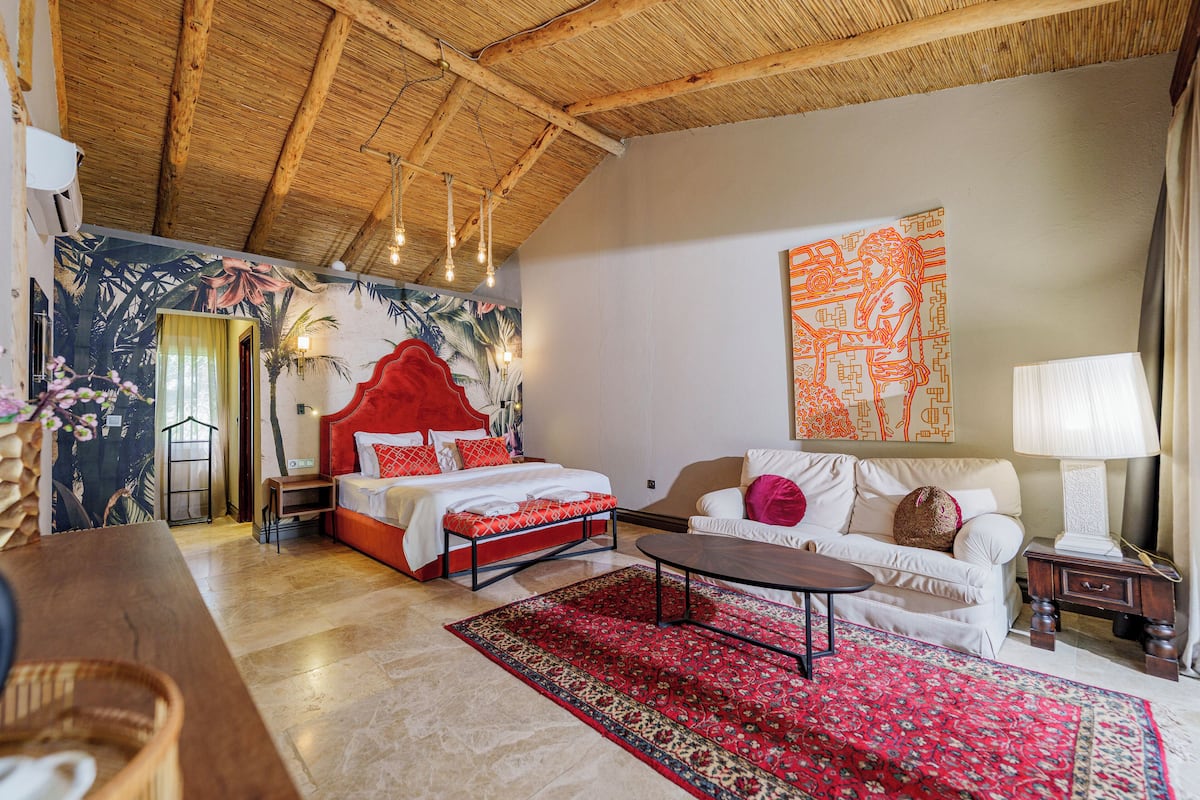 A room with a bed, a sofa, a coffee table, a red rug, and a wall with a tropical mural.
