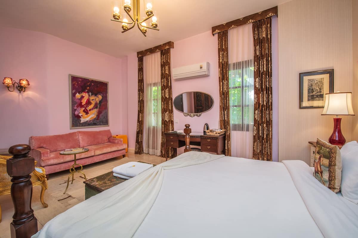 A hotel room with a bed, a pink sofa, a desk with a mirror, and a chandelier.