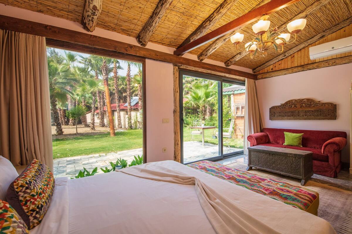 A bedroom with a large bed, a red sofa, and a view of a garden with palm trees.