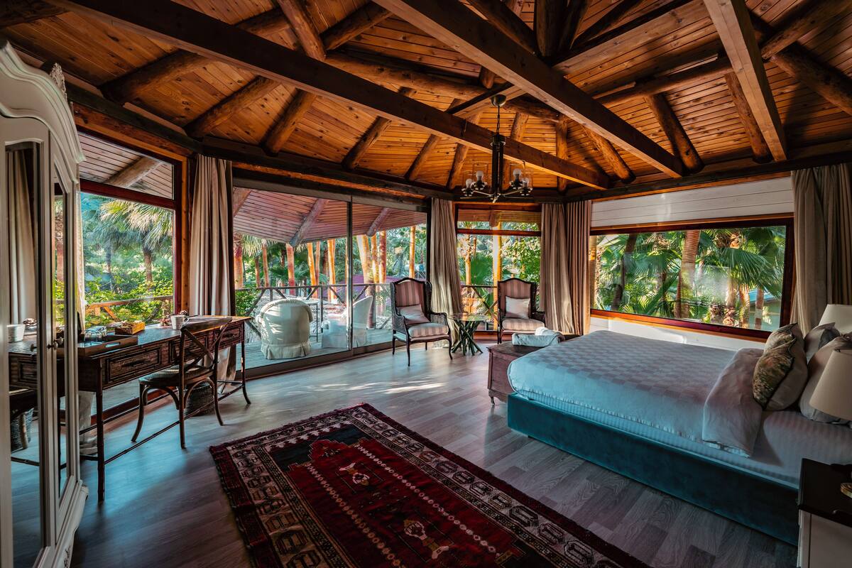 A spacious bedroom with a large bed, a desk, and a chair, with a view of lush greenery outside.