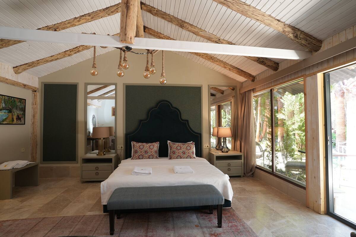 A spacious bedroom with a large bed, wooden ceiling beams, and a view of the outdoors.
