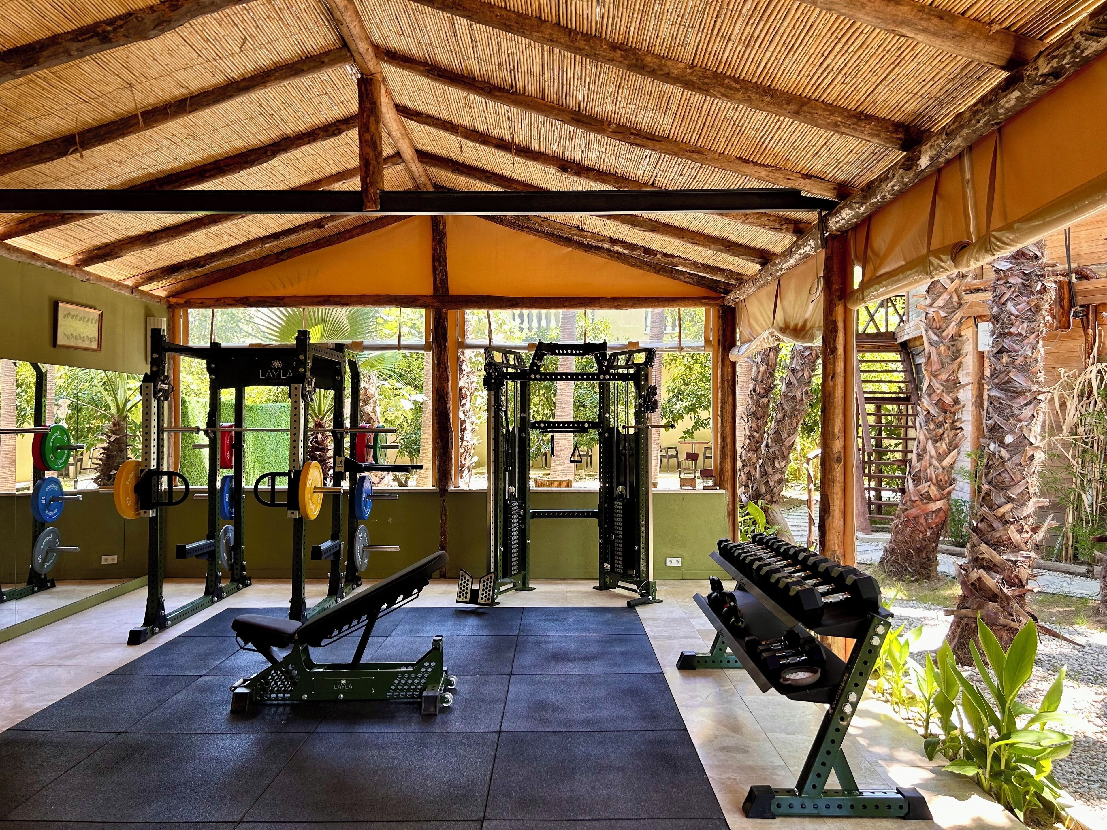 A covered gym with weightlifting equipment, a bench, and a view of greenery outside.