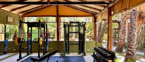 A covered gym with weightlifting equipment, a bench, and a view of greenery outside.
