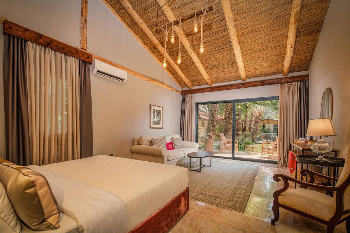 A hotel room with a large bed, a desk with a lamp, a chair, a sofa, and a view of the outdoors.