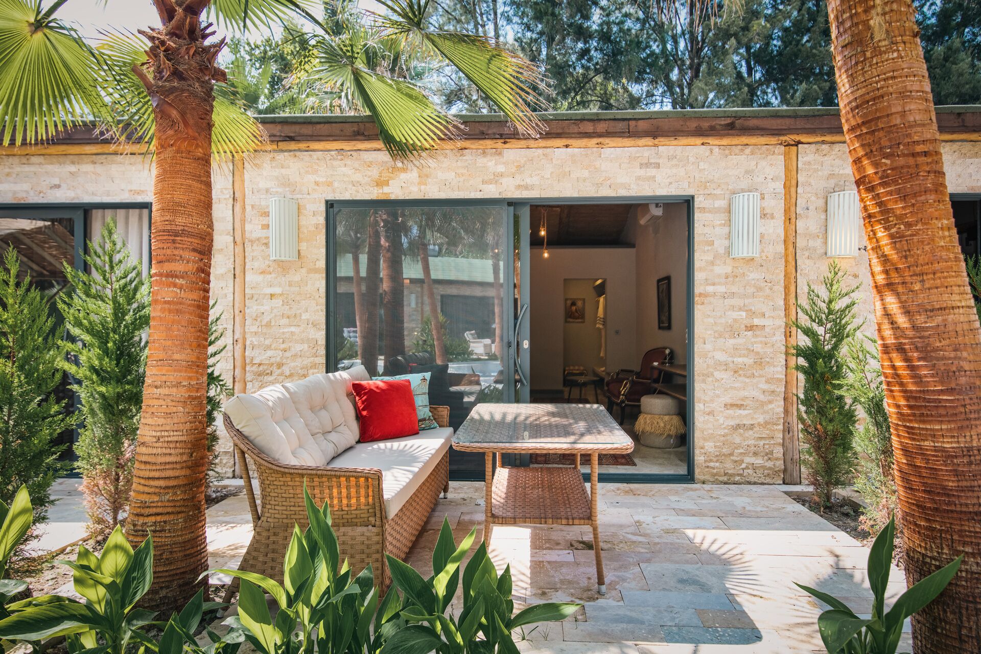 Un espace patio aménagé avec des meubles en osier, une porte vitrée donnant sur un espace intérieur, et une végétation luxuriante tout autour.