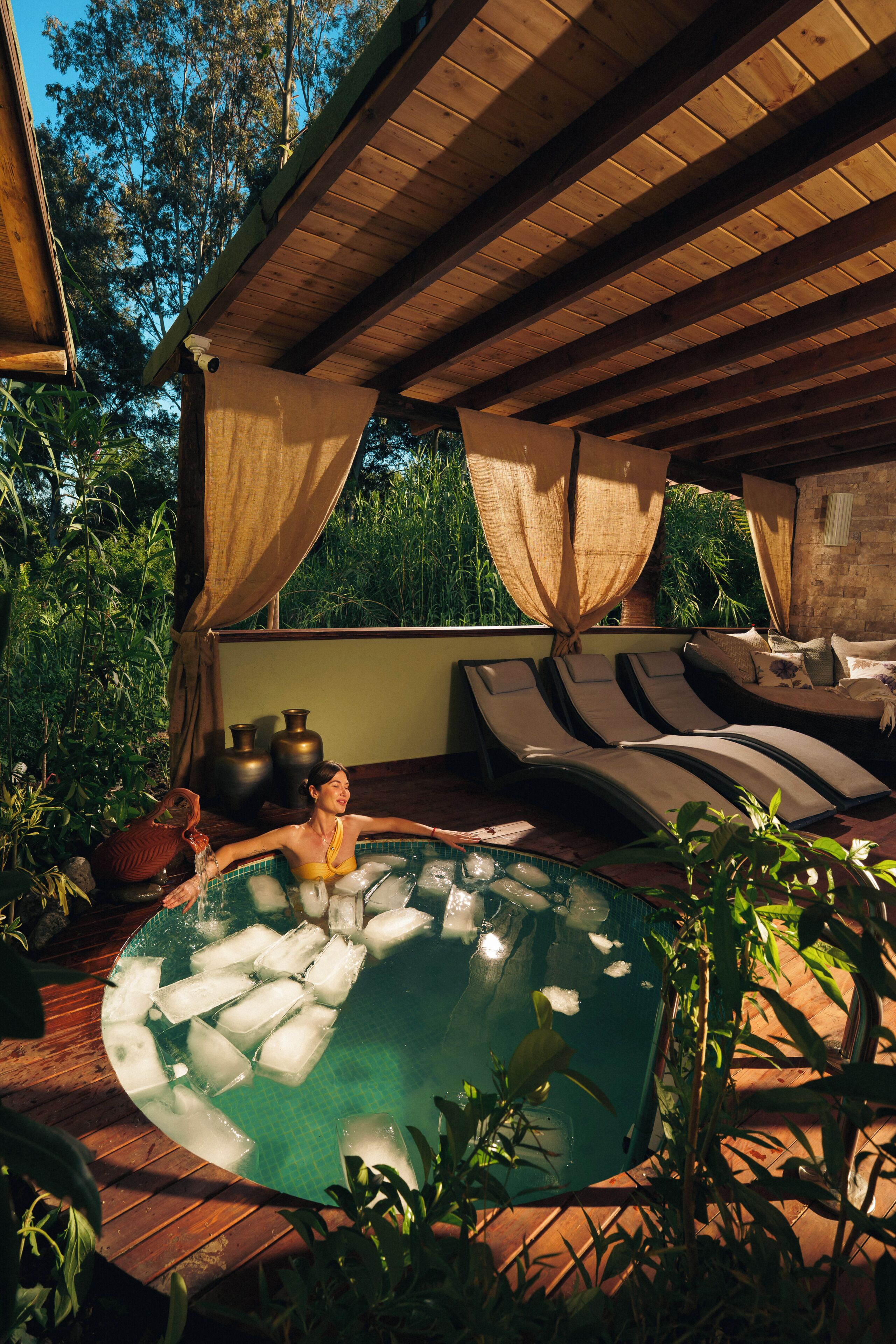 A pool area with lounge chairs and a hot tub surrounded by plants and a wooden pergola.