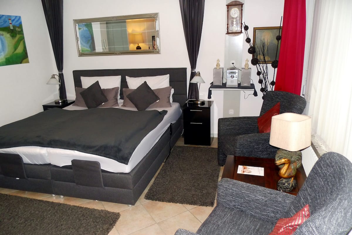 A hotel room with a large bed, a desk with a lamp, a chair, a clock, and a painting on the wall.
