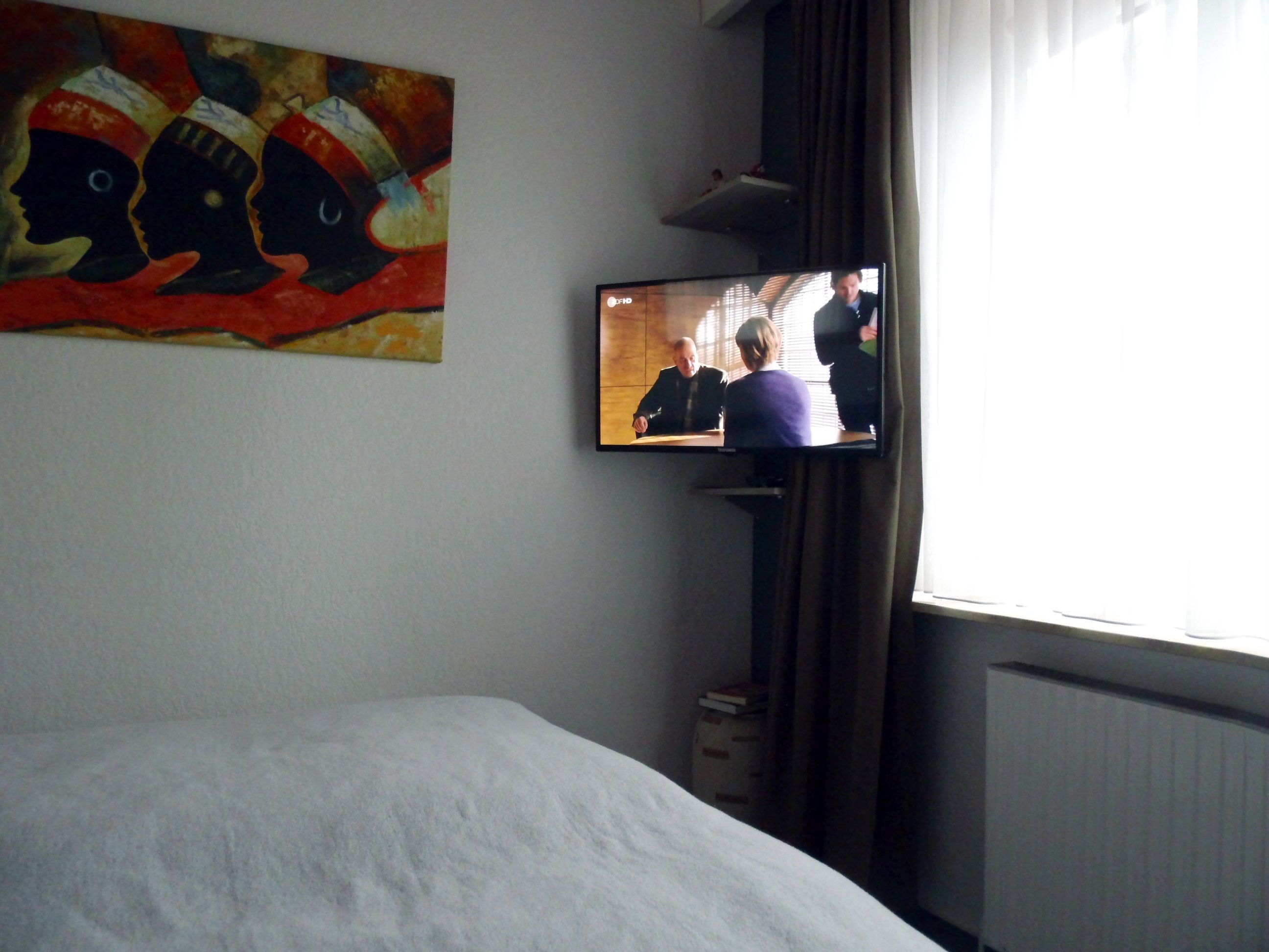 Habitación de hotel con una cama, televisor en la pared y un cuadro sobre la cama.