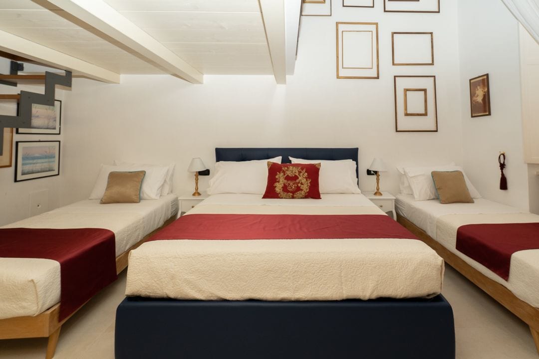 A hotel room with a large bed, two smaller beds, and a bench. There are framed pictures on the walls and a ceiling light fixture.