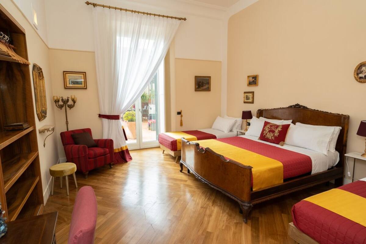 A hotel room with a large bed, two smaller beds, a red armchair, a wooden bookshelf, and a view through a glass door.