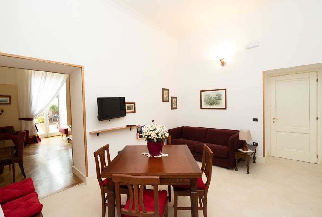 A living room with a wooden dining table, red chairs, a red sofa, a TV mounted on the wall, and a door.