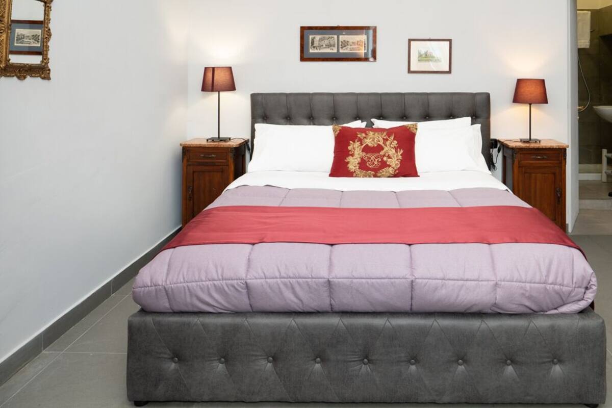 A neatly made bed with a grey upholstered headboard and footboard, white and red bedding, and two bedside tables with lamps.