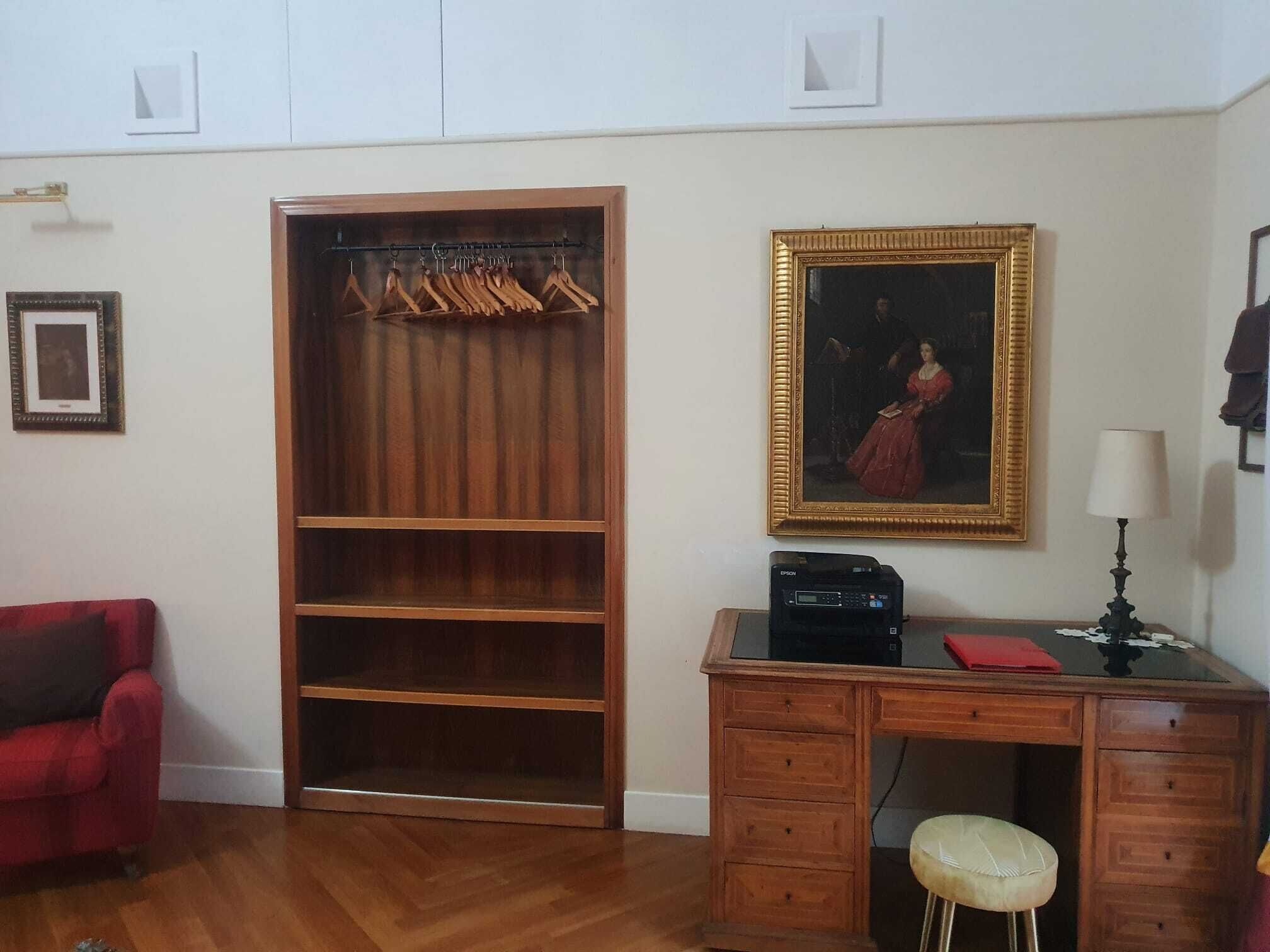 A room with a wooden wardrobe, a desk with drawers, a red sofa, a framed painting, and a lamp.