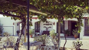 An outdoor patio with white metal chairs and tables, surrounded by potted plants and a white building with arches and a tiled floor. - Hotel Rural Molino la Boticaria (Marchena)