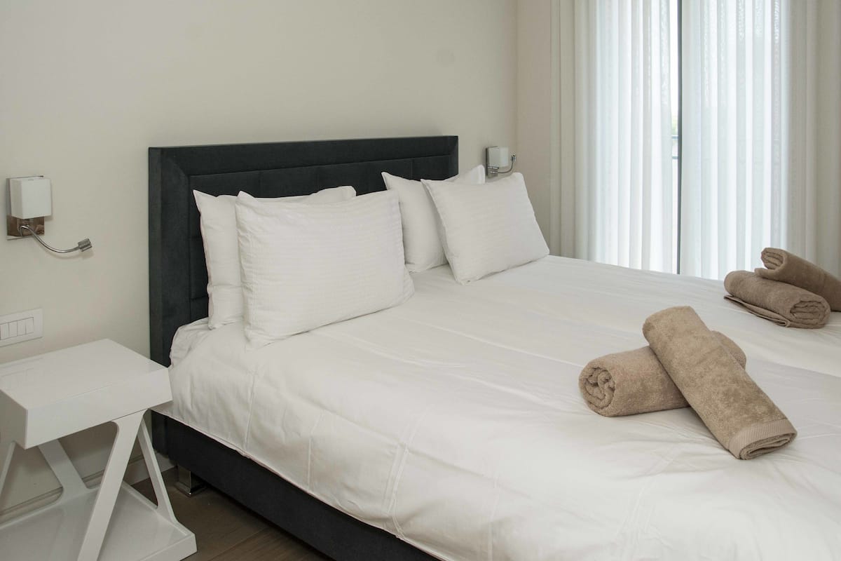 A neatly made bed with white linens, a dark headboard, and two rolled towels on the bed.