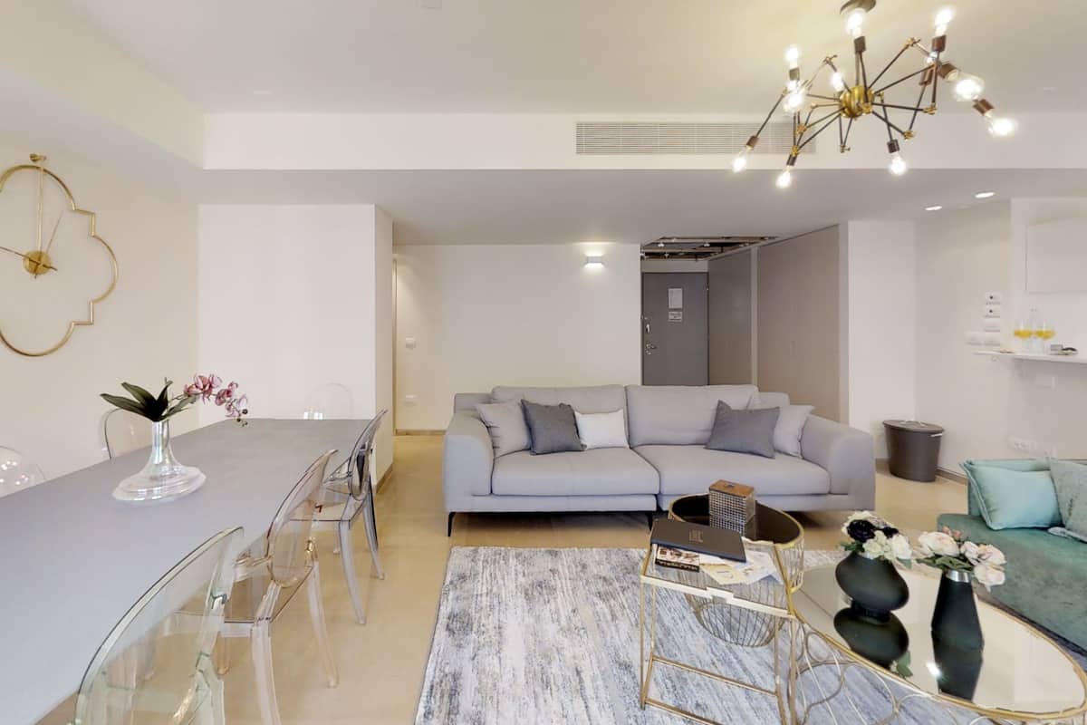 A modern living room with a grey sofa, a dining table with chairs, a chandelier, and a vase with flowers.