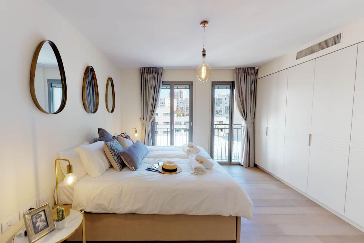 A bedroom with a bed, bedside lamps, a mirror, and a view of the outside.
