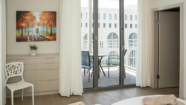 A hotel room with two beds, a painting, a chair, and a balcony with a view of buildings. - Rental Israel - Mamilla Residences (Jerusalem)