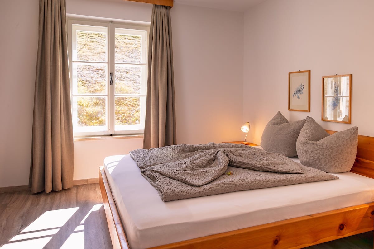 Un dormitorio con cama de madera, ventana con cortinas, lámpara de noche y dos cuadros en la pared.