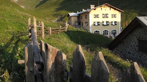 A two-story building with a yellow facade and green shutters, surrounded by a stone wall and wooden fence, set against a mountainous backdrop.