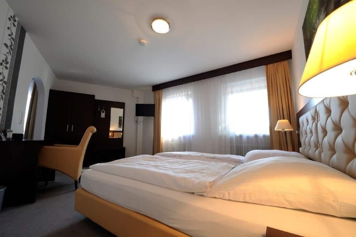 Quarto de hotel com cama, escrivaninha, cadeira e uma janela grande com cortinas.