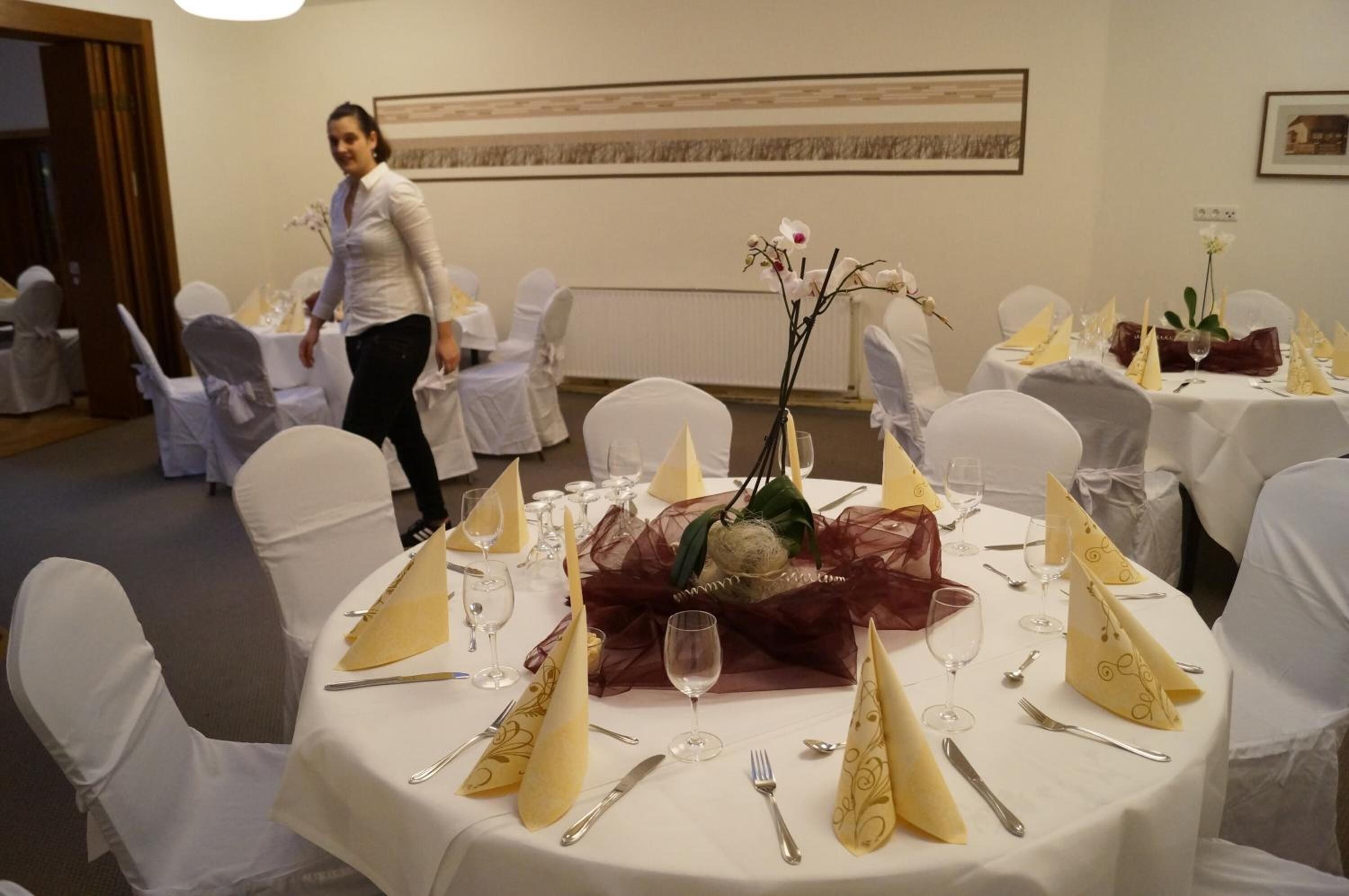 A banquet hall with round tables set for a formal event, featuring white tablecloths, gold napkins, and a central floral arrangement.