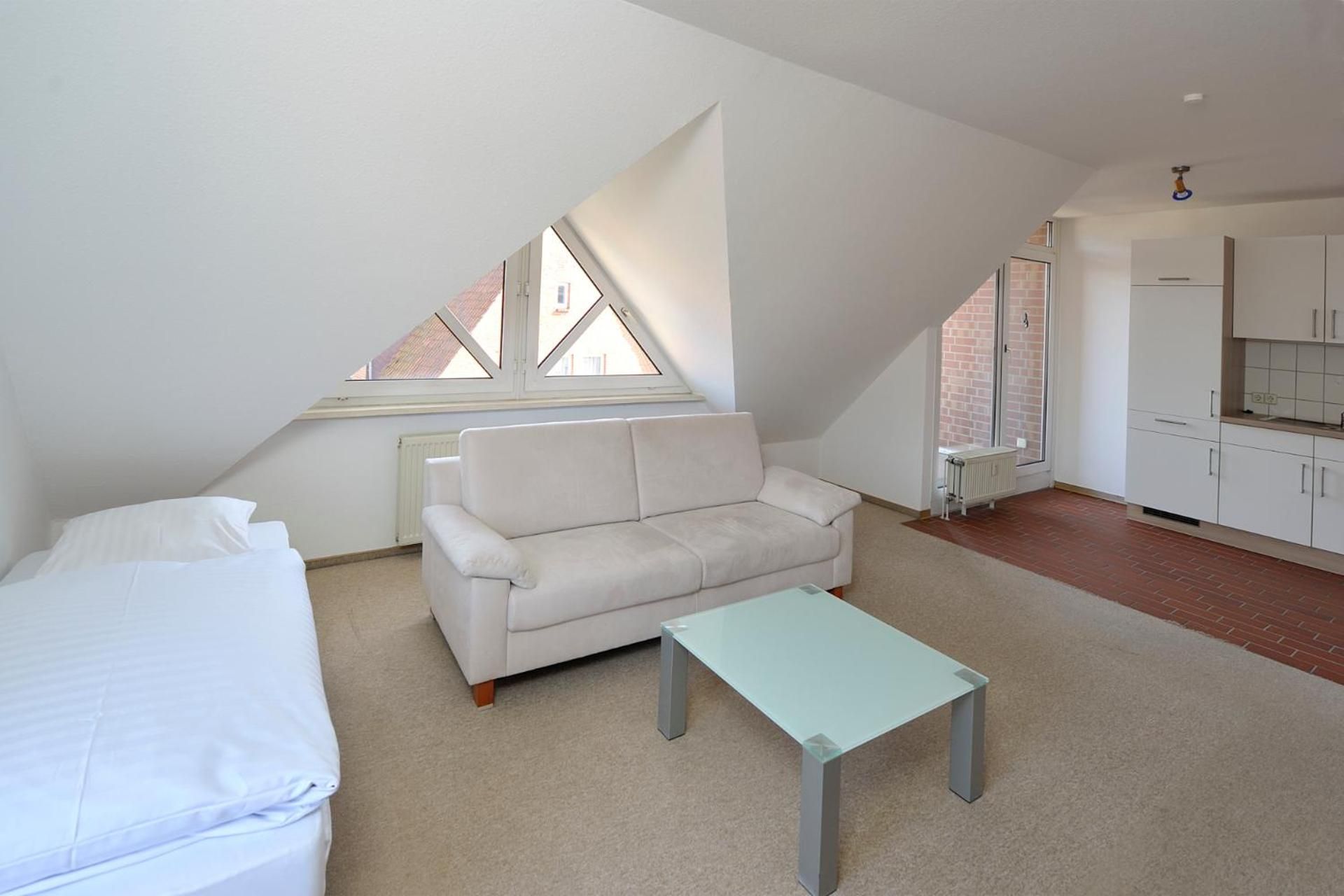 A modern, minimalistic room with a white sofa, a small table, and a bed. There are two windows providing natural light.