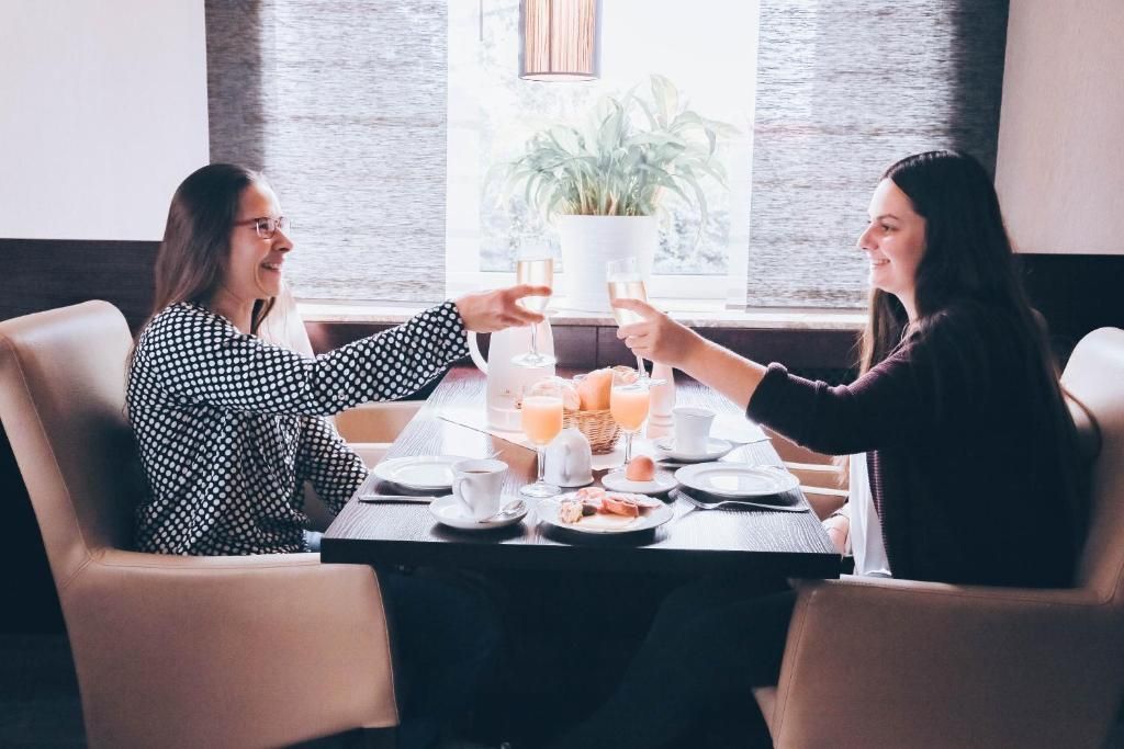 Two people toasting at a table with breakfast items and drinks, in a room with a window and a plant.