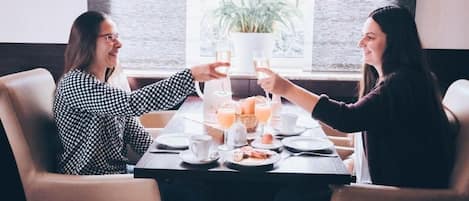 Two people toasting at a table with breakfast items and drinks, in a room with a window and a plant.