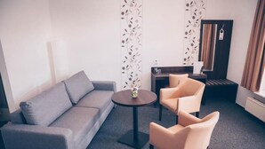 Une chambre d’hôtel avec un canapé gris, deux fauteuils, une petite table ronde avec un vase, une table d’appoint avec une bouteille et des verres, un miroir et un radiateur.
