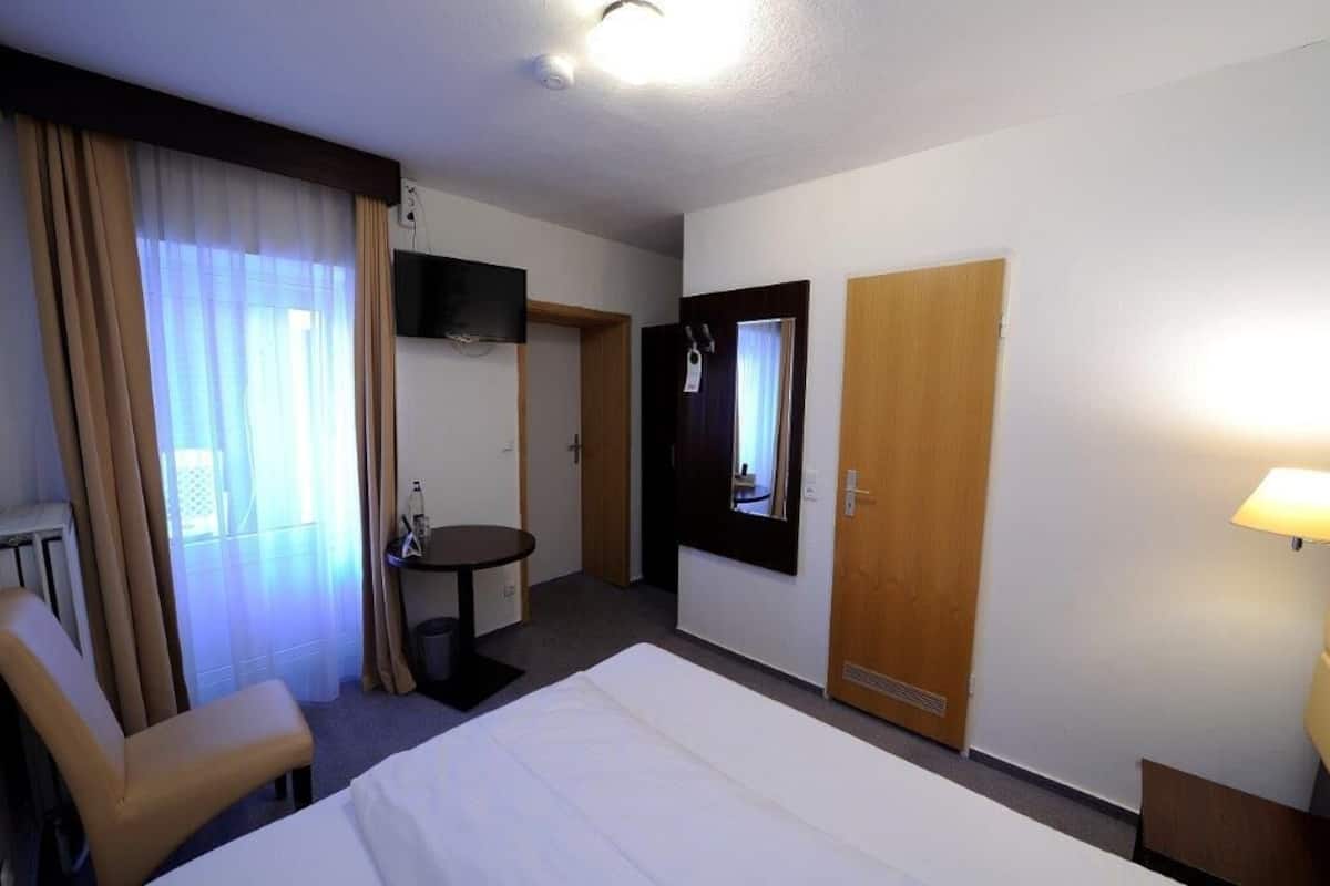 Quarto de hotel com uma cama, escrivaninha, cadeira, TV fixada na parede e porta com espelho.