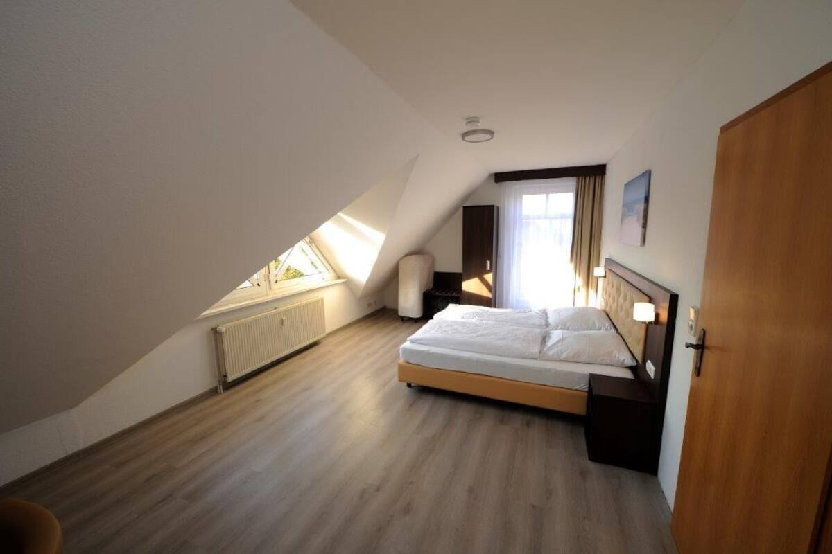 Quarto com teto inclinado, uma cama, mesa de cabeceira, janela com cortinas e radiador.