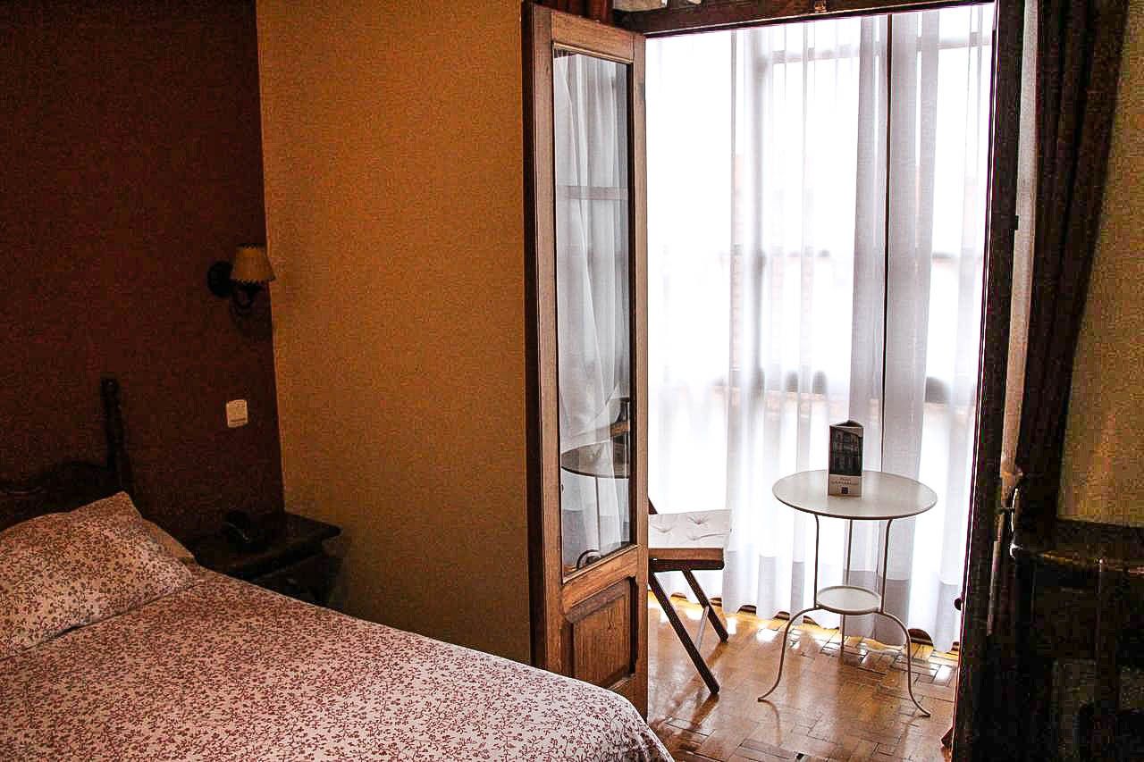 Een hotelkamer met een bed, een klein tafeltje met een lamp, en uitzicht naar buiten door een glazen deur.