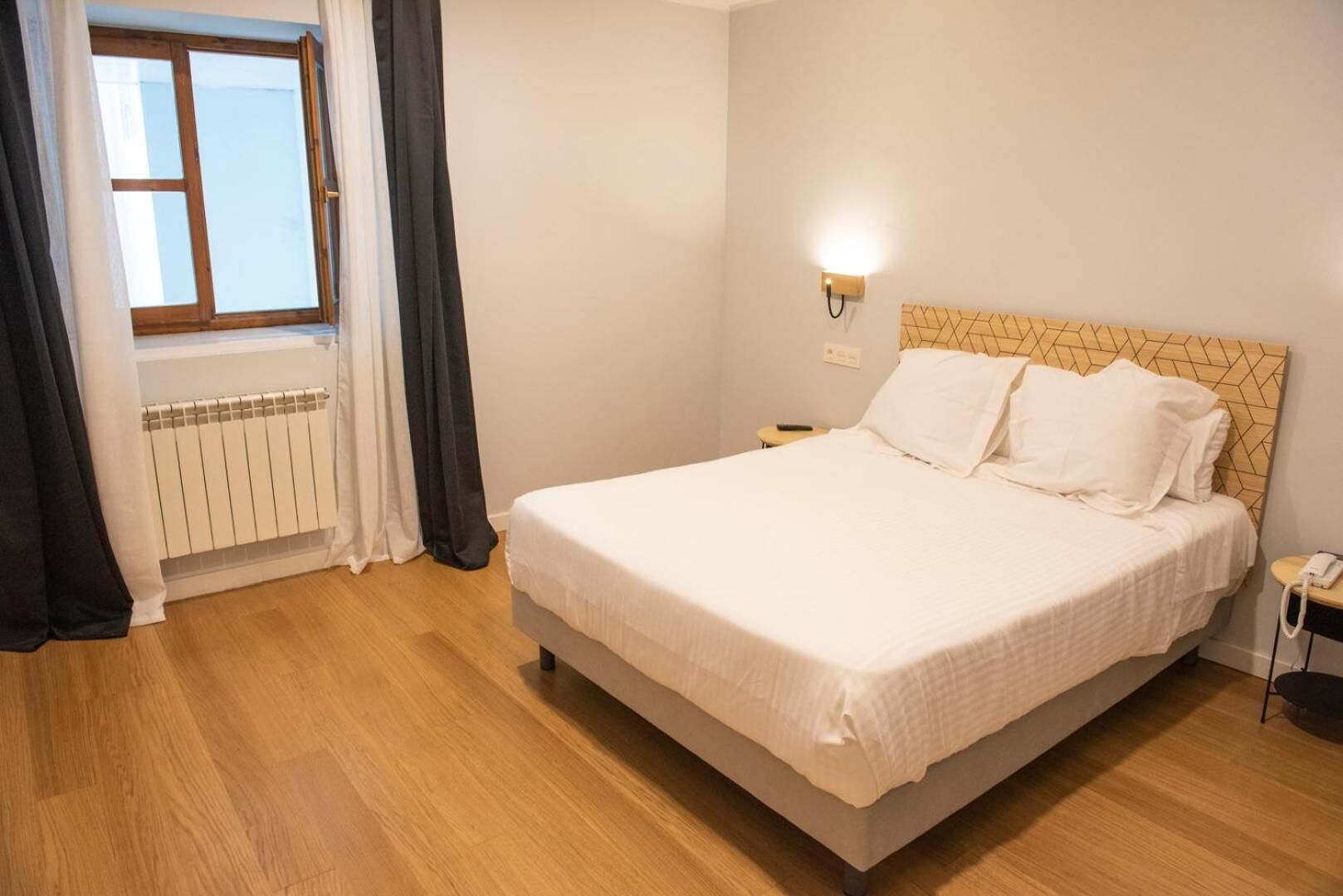A bedroom with a bed, a bedside table, a lamp, a window with curtains, and a radiator.